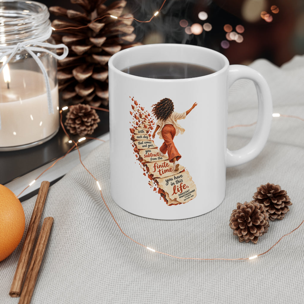 Finite Time white ceramic mug filled with a drink, handle on the right, beside a candle, pinecones and string lights.