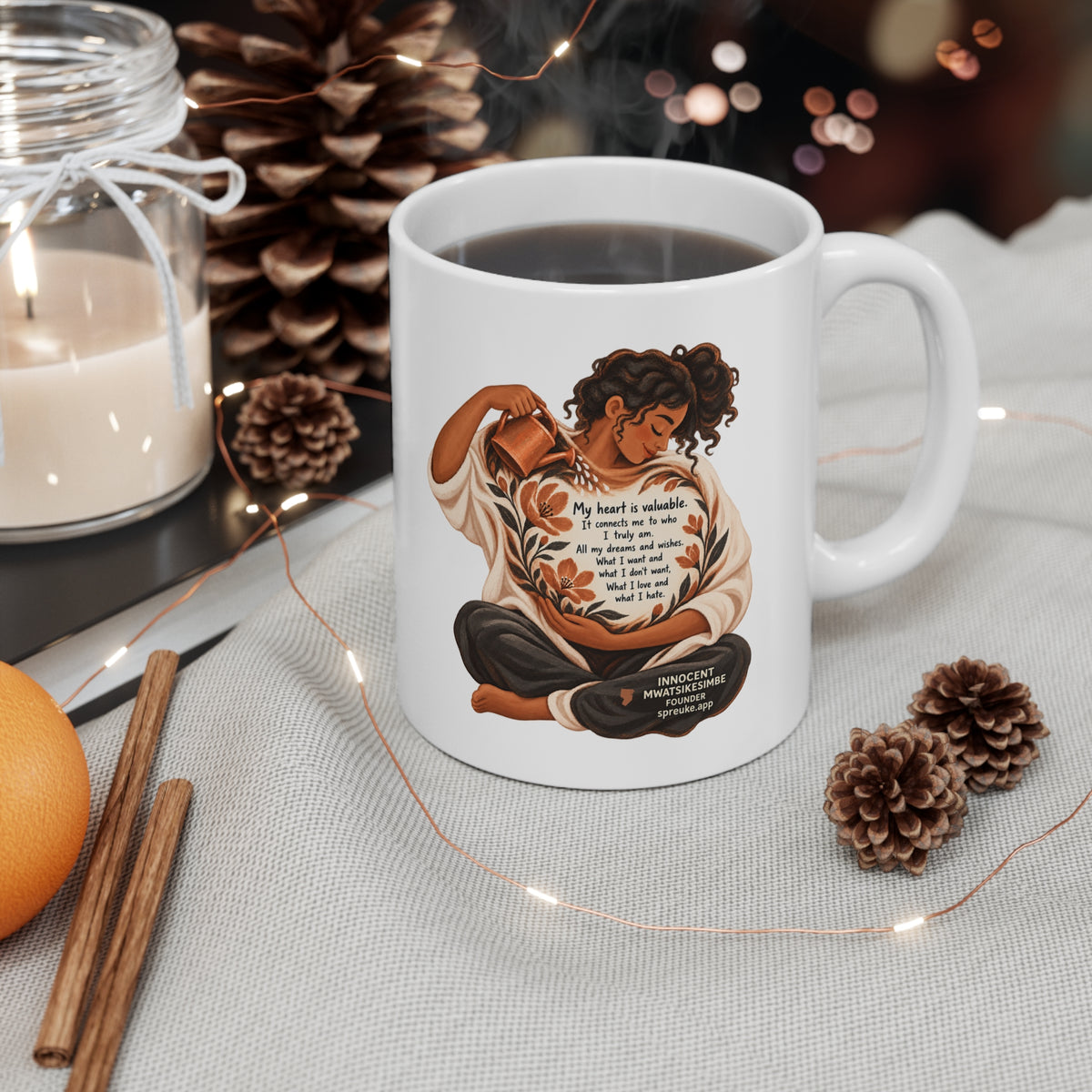 My Heart Is Valuable white ceramic mug filled with a drink, handle on the right, beside a candle, pinecones and string lights.
